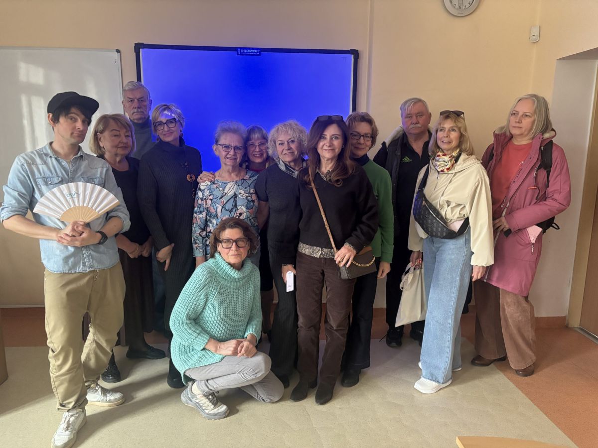 Na zdjęciu widać grupę kilkunastu dorosłych osób stojących razem w sali, prawdopodobnie szkolnej, na tle dużej tablicy multimedialnej. Uczestnicy są ubrani w różnorodne, codzienne stroje i patrzą w stronę aparatu, część z nich uśmiecha się, tworząc przyjazną, integracyjną atmosferę. Jedna osoba kuca z przodu, a reszta stoi blisko siebie, co sugeruje wspólne spotkanie lub zajęcia grupowe.