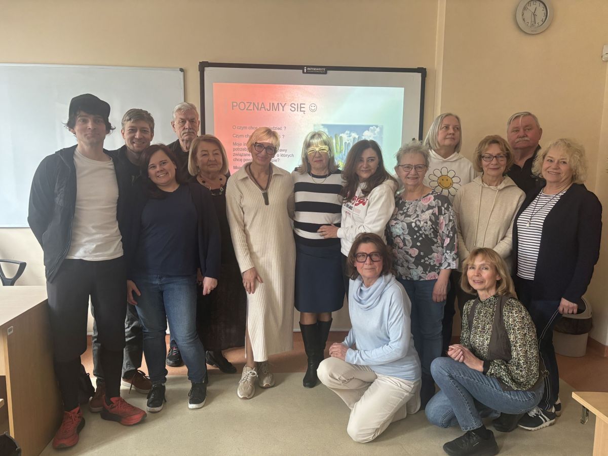 Grupa liczy 14 osób. Większość stoi w jednym rzędzie blisko siebie, a . Wszyscy są ustawieni tak, aby zmieścić się w kadrze wspólnego zdjęcia grupowego. Osoby są uśmiechnięte i wyglądają na zadowolone.