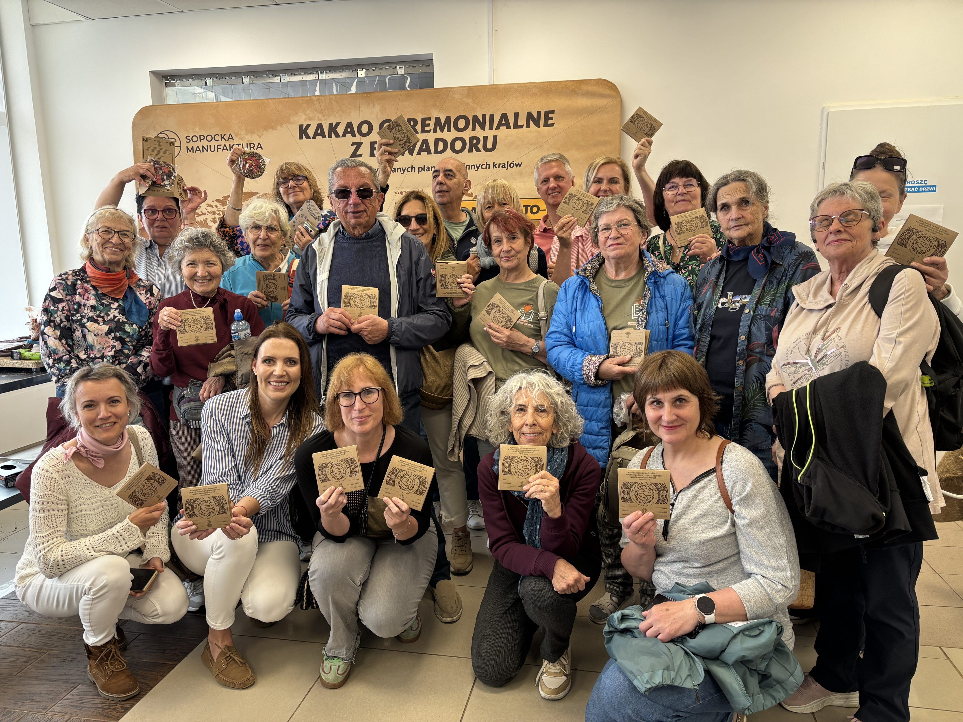 Na zdjęciu znajduje się duża grupa seniorów oraz kilku młodszych uczestników stojących i kucających we wnętrzu pracowni, wszyscy uśmiechają się i trzymają w dłoniach małe kwadratowe opakowania lub pamiątki związane z kakao ceremonialnym z Ekwadoru. W tle widoczny jest duży baner z napisem „Kakao ceremonialne z Ekwadoru”.