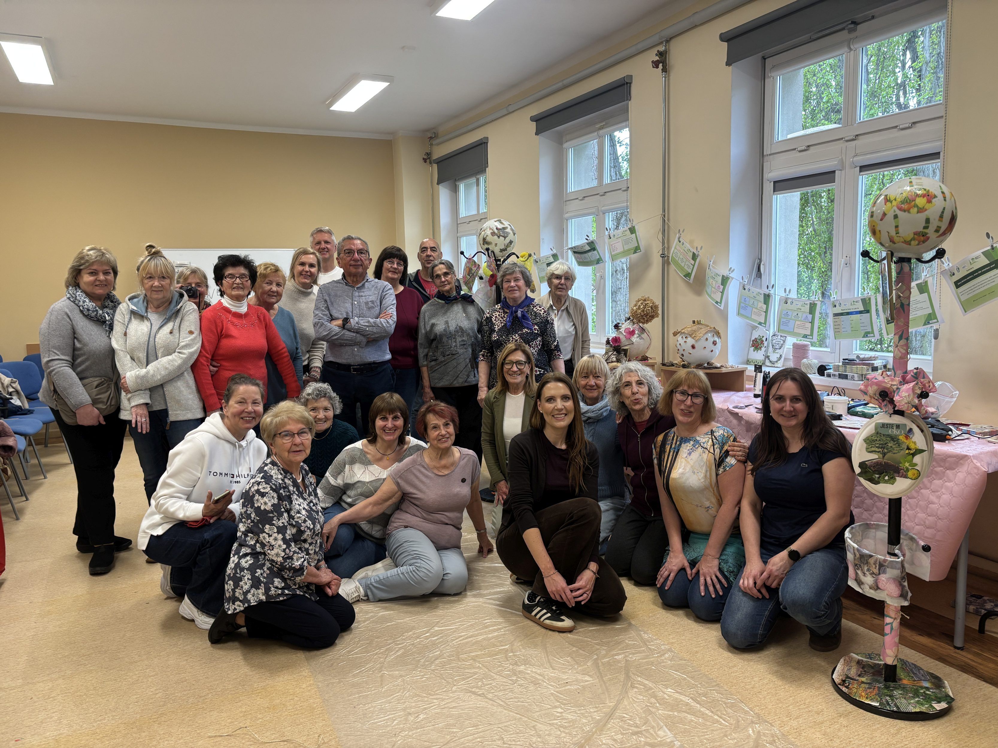 Na zdjęciu widoczna jest duża grupa seniorów i organizatorów pozujących razem w jasnej sali warsztatowej, część osób stoi, a część siedzi lub klęczy na podłodze, wszyscy uśmiechają się do aparatu. W tle znajdują się kolorowe prace ekologiczne i dekoracje wykonane podczas zajęć, zawieszone przy oknach i ustawione na stołach.