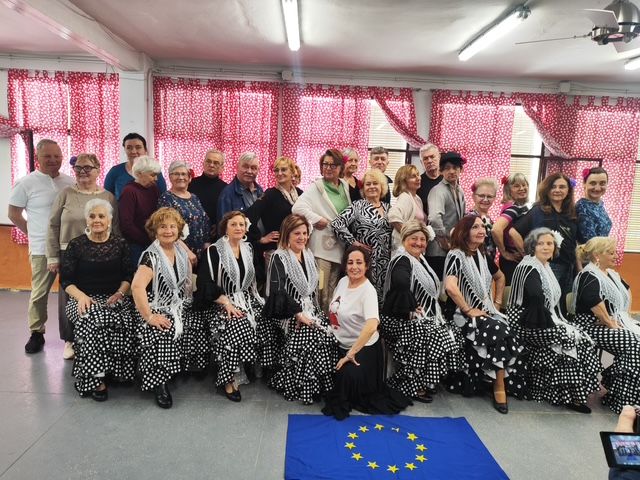 Duża grupa osób, głównie starszych kobiet i kilku mężczyzn, pozuje razem w sali, część siedzi z przodu w strojach przypominających taneczne – czarne sukienki w białe kropki z ozdobnymi szalami. Na podłodze przed nimi leży flaga Unii Europejskiej, a w tle widać czerwone zasłony w drobny wzór, co nadaje zdjęciu charakter pamiątkowy lub związany z występem
