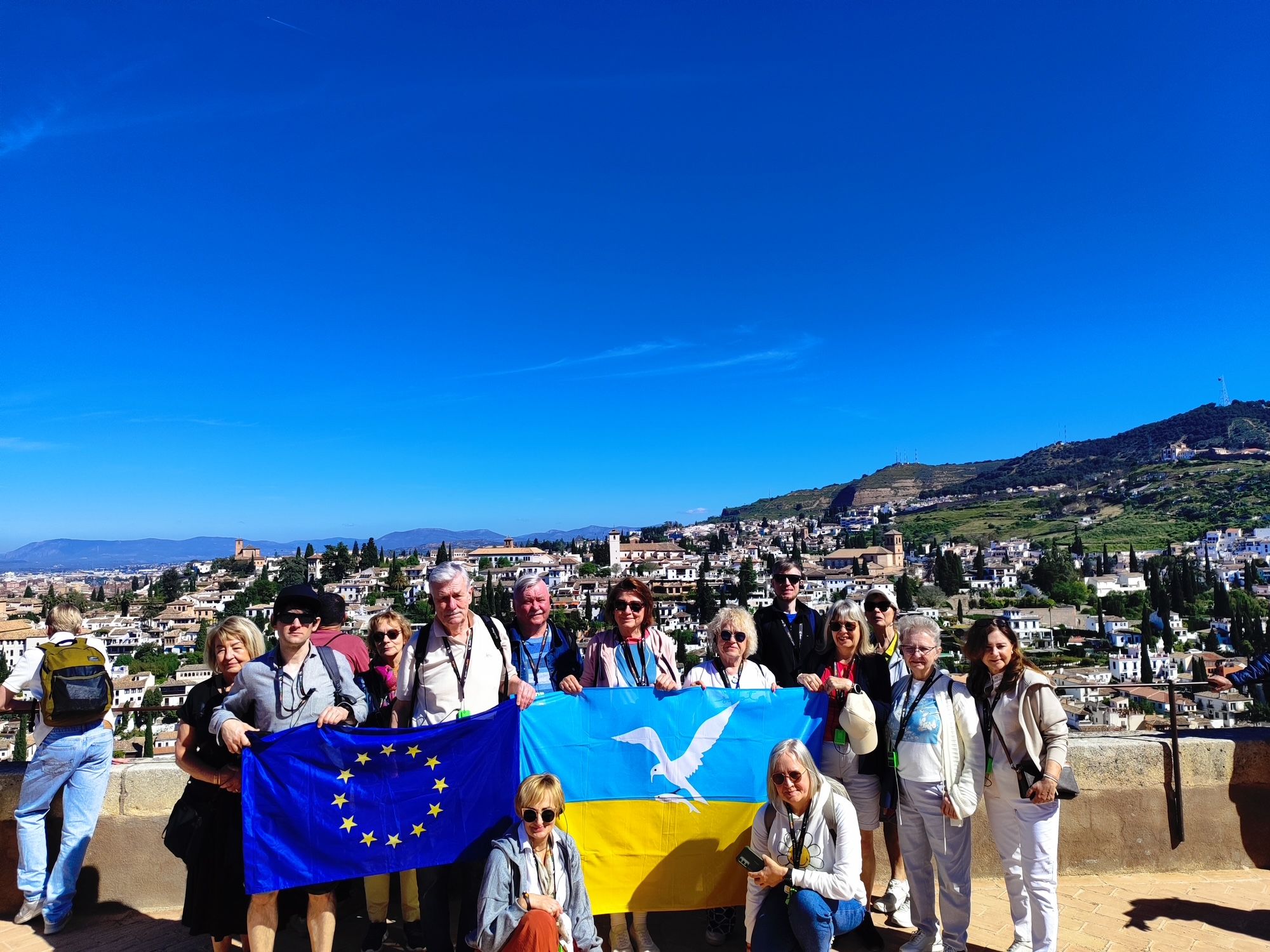Grupa kilkunastu turystów stoi na punkcie widokowym i pozuje do zdjęcia, trzymając flagę Unii Europejskiej oraz niebiesko-żółtą flagę z białym ptakiem. W tle widać rozległe, jasne miasto na wzgórzach pod bezchmurnym, intensywnie niebieskim niebem.